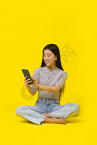 手机销售网站手持电话的亚洲女孩在地板上用手机发短信或买网上玩耍游戏 孤立于黄色背景 移动电话应用程序广告产品布置 复制空间 (续)背景