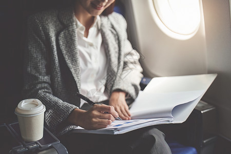 坐在纸飞机上一位成功的亚洲女商务人士或女企业家在飞机上穿着正式西装 坐在商务舱座位上 并在飞行期间使用带有文件的笔进行工作电脑管理人员裁剪航背景