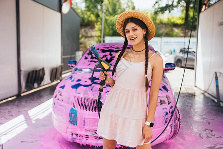 年轻的女青年水管站在一辆车前 车盖着粉色泡沫成人头发打扫工作女性黑发洗车短裤软管压力背景图片