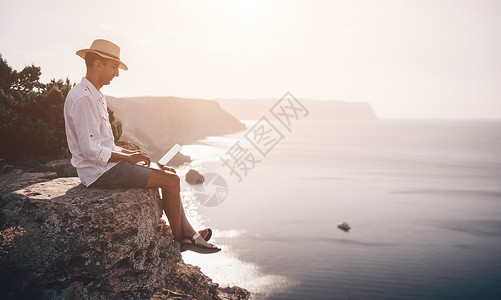 从横交错数字游牧者 自由职业者 假期远程工作 戴帽子的男人 一个带着笔记本电脑的商务人士 在日落时分坐在海边的岩石上 从远处在网上进行商背景