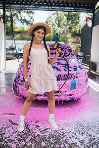 年轻的女青年水管站在一辆车前 车盖着粉色泡沫工作软管女性头发洗车压力短裤肥皂辫子女孩背景图片