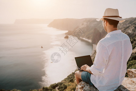 从横交错数字游牧者 自由职业者 假期远程工作 戴帽子的男人 一个带着笔记本电脑的商务人士 在日落时分坐在海边的岩石上 从远处在网上进行商背景