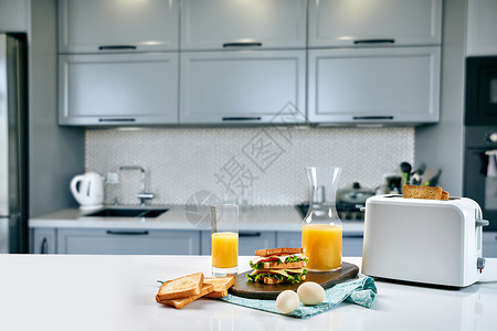 地蛋餐桌上有橙汁和三明治的白色电动面包面包机 在舒适的厨房里吃早餐电气橙子机器花朵水果炙烤工具木头器具制作者背景