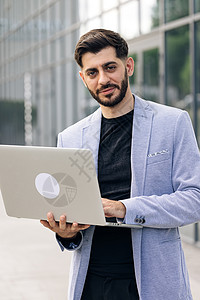 留着胡须的年轻成功商务人士自由职业者正站在现代办公室附近 看着镜头微笑 男人正在他的笔记本电脑上工作背景图片