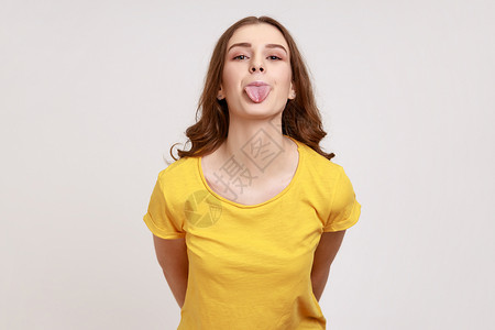 逗逼表情穿着黄色T恤的年轻女性 带着幼稚面部表情和舌头露出笑柄 看着镜头 看相片背景