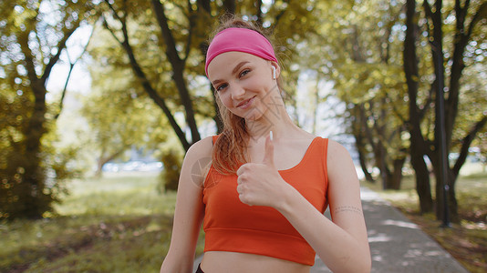 运动式微笑跑跑步女孩的肖像 看着镜头 举起拇指 同意一些事情运动员爱好跑步有氧运动训练耐力身体马尾辫活动音乐背景
