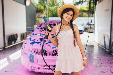 年轻的女青年水管站在一辆车前 车盖着粉色泡沫工作肥皂打扫女性女孩短裤软管喷射压力头发背景图片