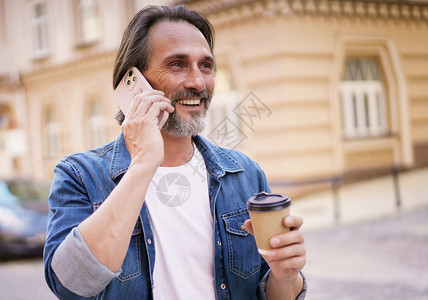 快乐的中年男子 留着灰色胡子 拿着一次性纸杯里的咖啡 站在户外 身穿牛仔裤衬衫 穿着牛仔裤 自由职业者旅行人背景图片