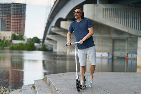 不要去河边在户外工作后 骑着滑板车英俊时尚的中年男子 留着灰色的胡子 站在河边的城镇桥下 背景是城市 旅行 生活方式概念背景