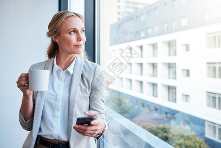 当头脑清晰时 任务就变得清晰了 一位成熟的女商务人士一边使用智能手机一边看着窗外 在现代办公室里喝咖啡背景