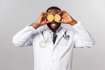 健康护理 健康饮食和疾病概念 快乐的非洲美国男医生穿白色大衣 激发儿童躺在医务室的情绪 解释病人多吃维他命 用橙子盯着眼睛社交药背景图片