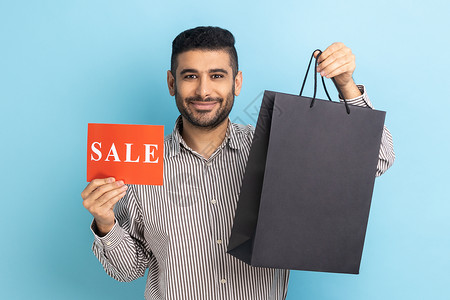 范冰冰男人装男人装着购物袋和卡片 手持销售标记 看着相机 笑声营销喜悦拉丁购物者商务展示蓝色人士黑色价格背景