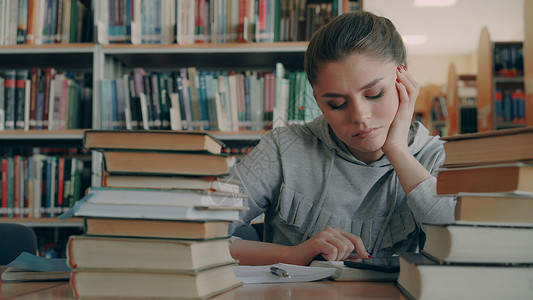 天主教妇女坐在大学图书馆的桌子上 周围有一堆藏着数字平板电脑的书籍 她疲惫不堪 精疲力尽教育考试写作学生药片图书女士学习教科书女背景图片