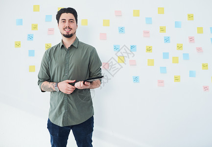 一位年轻商务人士的肖像 他用数字平板电脑站在墙上 办公室里贴着纸条 站着一面墙背景图片