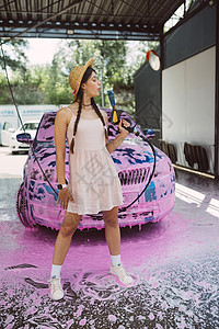 年轻的女青年水管站在一辆车前 车盖着粉色泡沫洗车打扫肥皂喷射辫子压力女性黑发短裤头发背景图片