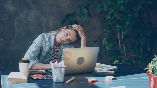 年轻的金发女人正坐在办公室里用电脑工作 她在一整天的工作后感到疲倦背景图片