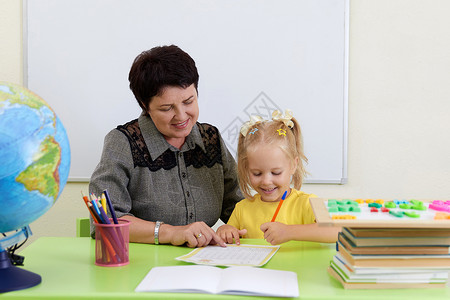 坐在桌边的成熟老师教小女孩写字 教她写字学校女士班级导师幼儿园微笑桌子女性工作女孩背景图片
