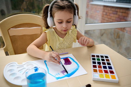 技能大师戴耳机的可爱学龄前女孩 小艺术家画画 用水彩颜料 爱好 美术大师班背景