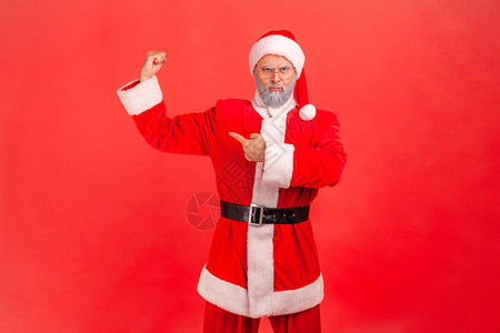 我很坚强 我可以做任何事 留着灰色胡子的老人穿着圣诞老人服装站着指着并展示他强壮的二头肌背景图片