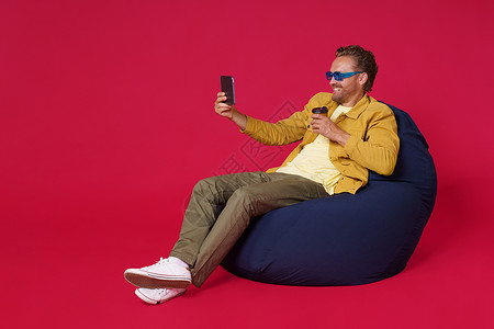 快乐英俊的年轻人 30 多岁使用数字平板电脑从纸杯喝咖啡自拍穿着休闲牛仔装坐在袋椅上隔离在红色背景 咖啡店经营理念商业眼镜杯子衣背景图片