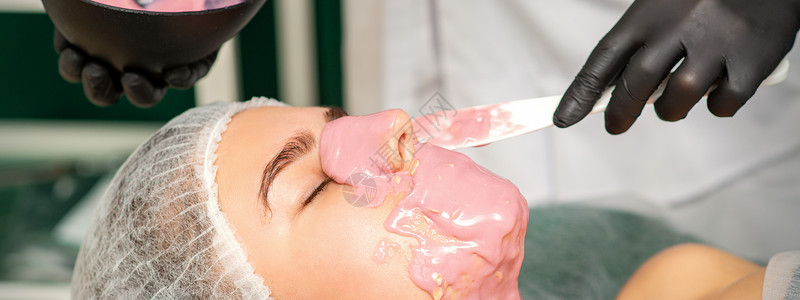 皮肤光滑的脸美容师在美容院对一位年轻女子的脸部 戴上悬浮面罩女孩女性温泉皮肤塑化奢华呵护美容沙龙程序背景