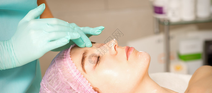 面部松弛戴着手套的美容师在女性脸上涂上带去角质霜的保湿面膜 面部美容治疗 面部护理程序治愈者诊所病人皮肤酵素美丽女士医生奶油去角质背景