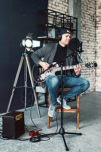 一个男歌手坐在一个椅子上 耳机里坐着一小凳子 吉他在家庭工作室录音演员男性歌曲音乐家创造力电缆人声独创性嗓音抗震背景图片