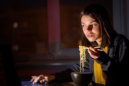 女孩在笔记本电脑上吃面和看录像命令外卖电视节目视频女人工作互联网办公室杯子失眠背景图片