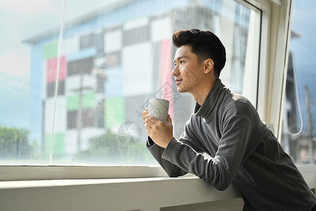 坐在办公室窗户旁边 俯视着城市景色 享受清晨咖啡的可爱英俊的亚洲男子图片