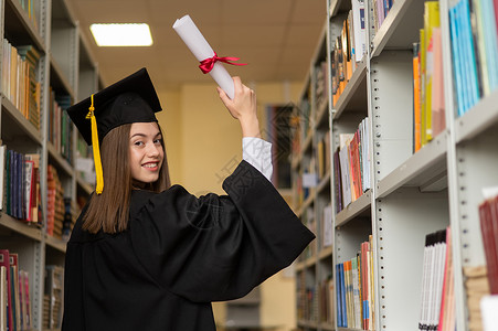 证书架子穿着毕业礼服的快乐年轻女子 在图书馆持有文凭流苏帽子研究生学士大学女孩女性法律知识庆典背景