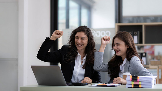 办公室好素材两位年轻的亚洲女商务人士在现代办公室里用笔记本电脑开心地微笑着 表现出工作成功的喜悦表情职员会议商务学生团队女性快乐手臂庆典人士背景
