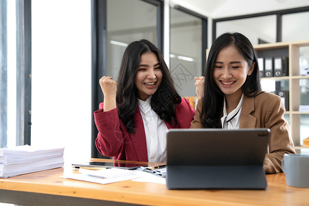 两位年轻的亚洲女商务人士在现代办公室里用笔记本电脑开心地微笑着 表现出工作成功的喜悦表情手臂快乐会议人士女性两个人职业职员女士商背景