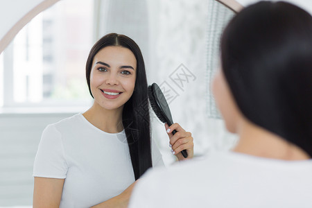 光泽度高一位美丽的年轻女士站在镜子里看着自己 用梳子梳头发 她把头发梳了一下衬衫女性发型刷子浴室成人派对护理黑发微笑背景