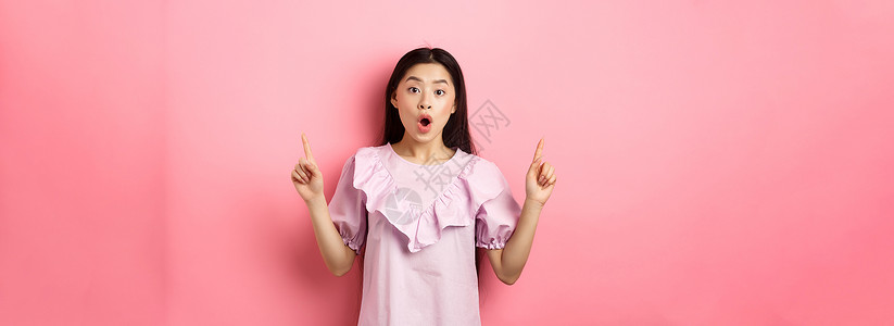 兴奋的亚洲青少年女孩举起手指 说哇和展示宣传协议 站在粉红背景上穿着着衣服情感广告化妆品促销学生女士投标女性工作室购物背景图片