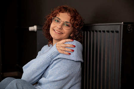 笑着的女孩坐在热散热器旁边 靠着她倾斜生态房间回暖持有暖气眼镜活力系统安装热能背景图片
