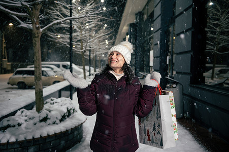 可爱的女士拿着购物袋 捕捉雪花 走在由节日园林照亮的雪街上背景图片