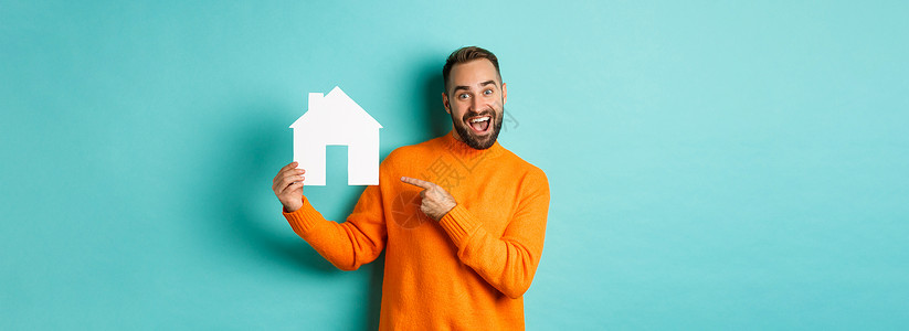 地产概念 快乐的男子用手指对着纸牌房子和微笑 站立在浅蓝背景上标识男人横幅胡子毛衣购物抵押情感黑发信用背景图片