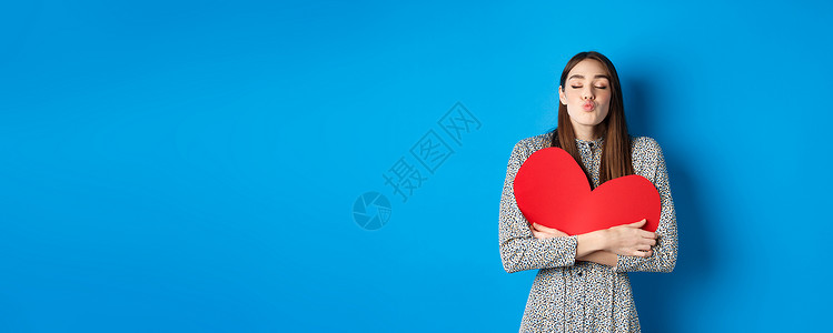 浪漫的美丽美女闭上眼和嘴唇亲吻 握着大红心胸切开 亲吻你 站在蓝背景上工作室灵魂纪念日周年惊喜伙伴庆典女性女朋友化妆品背景图片