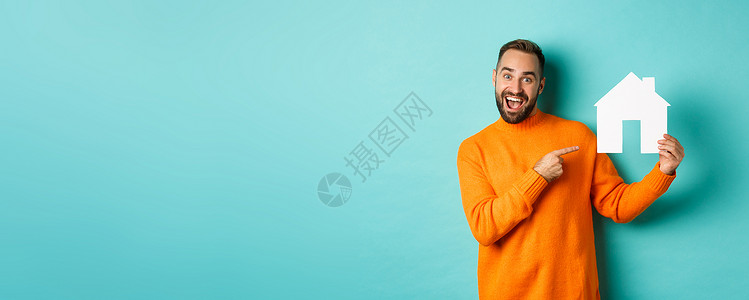 地产概念 快乐的男子用手指对着纸牌房子和微笑 站立在浅蓝背景上广告潮人男人信用抵押胡须公寓市场黑发房地产背景图片