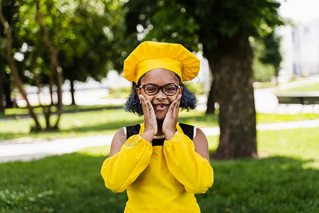 身着厨师帽和黄色围裙制服的非洲黑人儿童厨艺女孩握着脸颊并感到惊讶 为咖啡馆或餐馆制作创意广告烹饪黑色乐趣帽子食物孩子童年微笑孩子背景图片