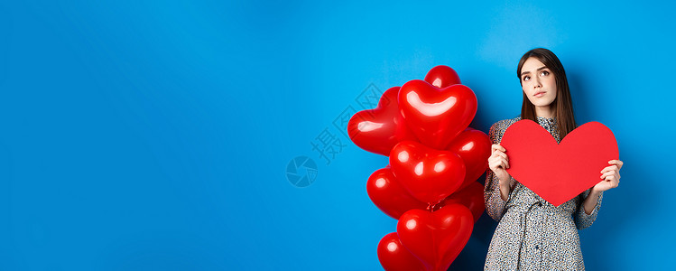 大红色气球情人节 梦幻般的漂亮女士穿着裙子 拿着大红色的心形剪纸 寻找真爱 抬头沉思 站在蓝色背景的假日气球旁微笑庆典工作室女朋友礼物情感背景