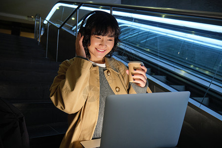 舱外作业美丽的年轻女学生 韩国妇女坐在楼梯上 观看耳机笔记本电脑的视频 喝外送咖啡 在公共场所远程办公工作;以及背景