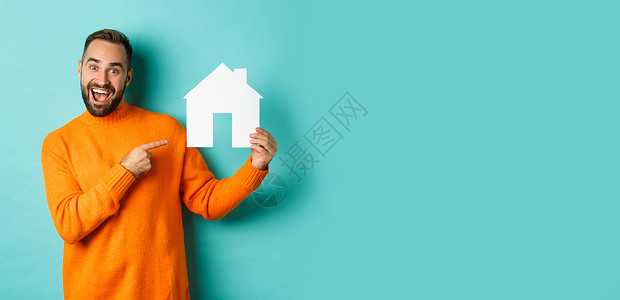 地产概念 快乐的男子用手指对着纸牌房子和微笑 站立在浅蓝背景上毛衣广告公寓房地产促销信用男人情感购物潮人背景图片