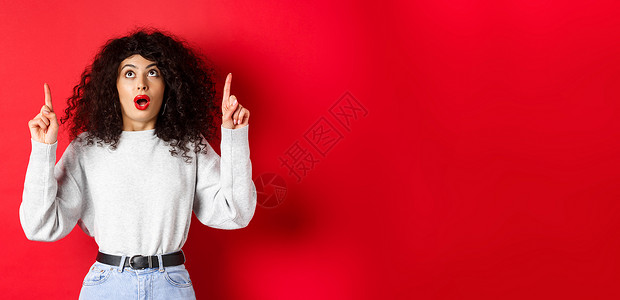 令人印象深刻的欧洲女性模特身着临时服装 用下巴检查宣传方案 抬头看和指手指 展示标志 站在红色背景上站立促销购物嘴唇女士发型卷曲背景图片