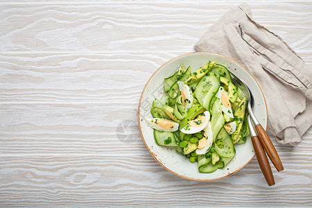 白绿色菜单健康的绿鳄梨沙拉碗 配有煮鸡蛋 切片黄瓜 豆子 橄榄油和陶瓷板顶端的草药 白木制餐桌背景背景