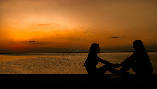 两个太阳素材两个漂亮的女孩坐在沙发上牵着手 在日落和橙色天空的海边玩得开心 暑假和背包旅行的概念是背景