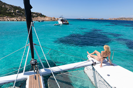 在意大利撒丁岛马达莱纳群岛的 Spargi 岛上 坐在豪华双体船上的女人在夏季航行中放松 靠近完美的白色沙滩 太阳 血管背景