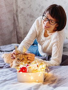 可爱的姜黄色猫 有着由装饰性羽毛制成的亮金色耳朵 女人在玩她毛茸茸的宠物和装有圣诞装饰品的盒子 新年前舒适的家 十二月 毛皮背景图片