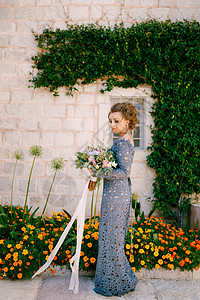 蓝色瓜叶菊一位身穿蓝色裙子 拿着花束的新娘站在一栋房子的墙上 墙上挂着绿色的藤本植物和橙色的花 特写背景