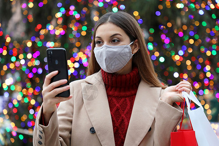 哦买噶快乐的年轻女子带着面罩 手拿着购物袋 在户外用智能手机买圣诞礼品吧 - 哦 不!背景